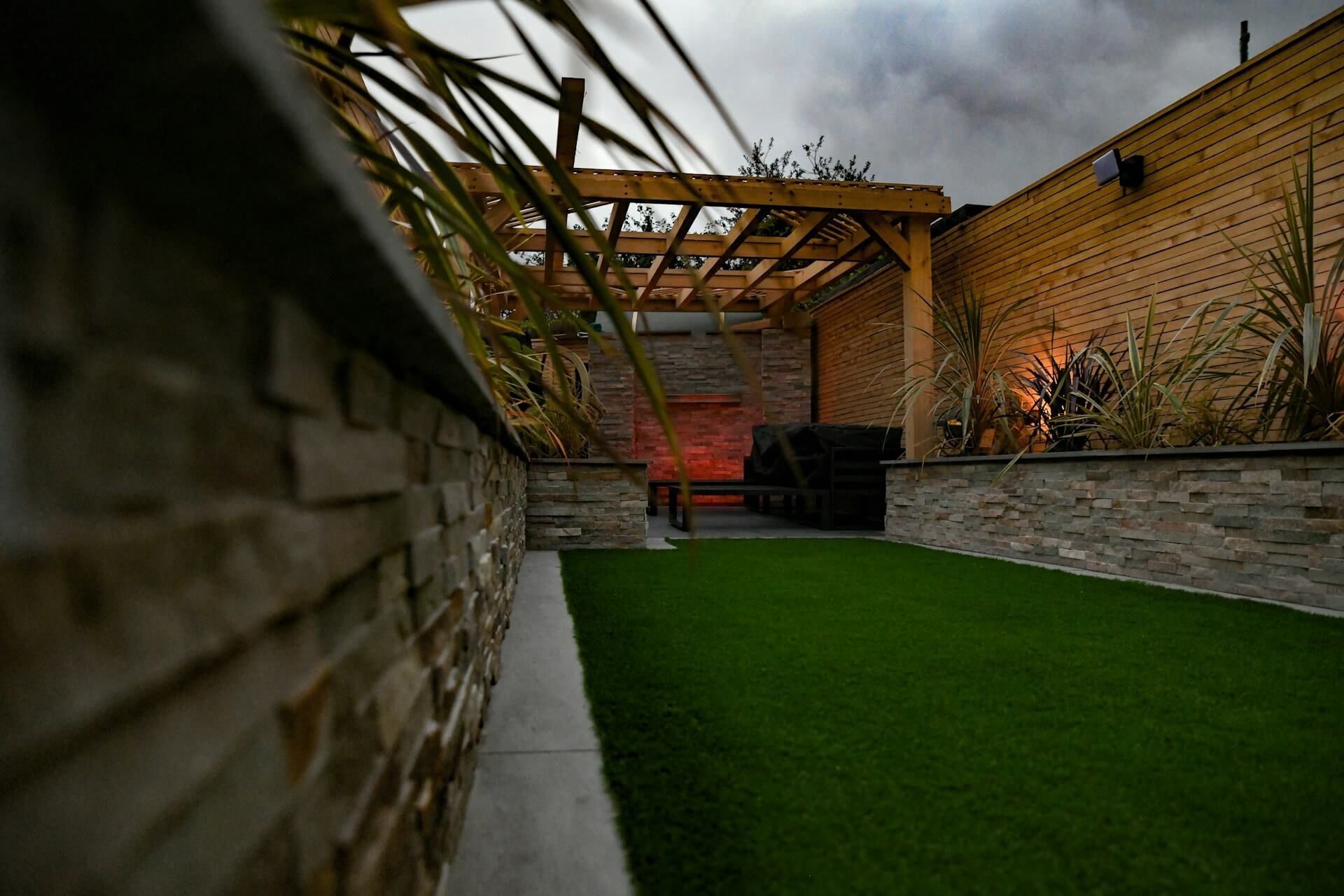 Modern wooden garden pergola with a seating area, stone walls, and decorative lighting, set on artificial grass.